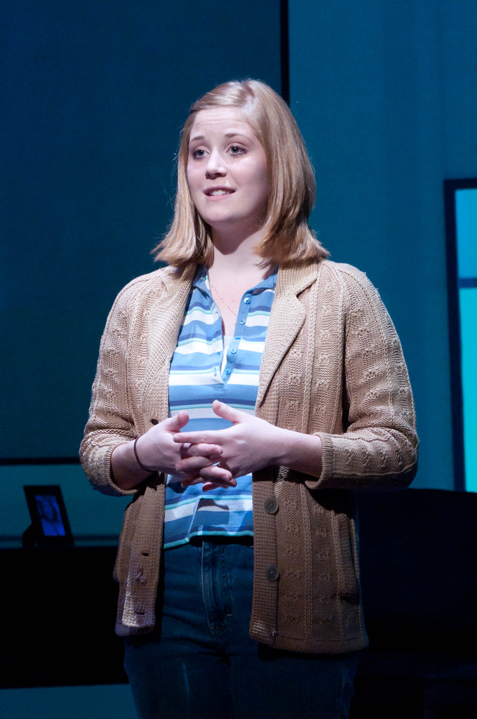 A Bethany Lutheran College student stands on stage wearing a tan cardigan and striped shirt, hands clasped in front of her.