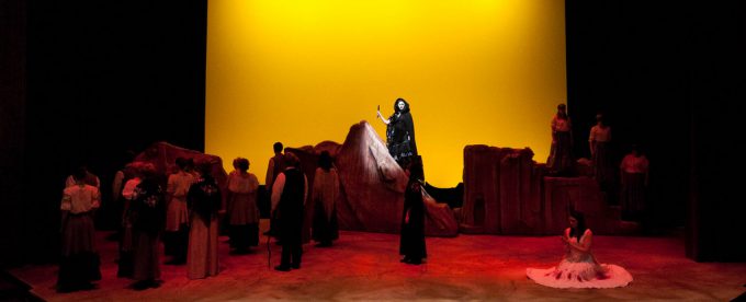 Bethany Lutheran College students perform in a theatrical production featuring actors in period costumes onstage, with a vibrant yellow backdrop and rocky set pieces enhancing the scene.