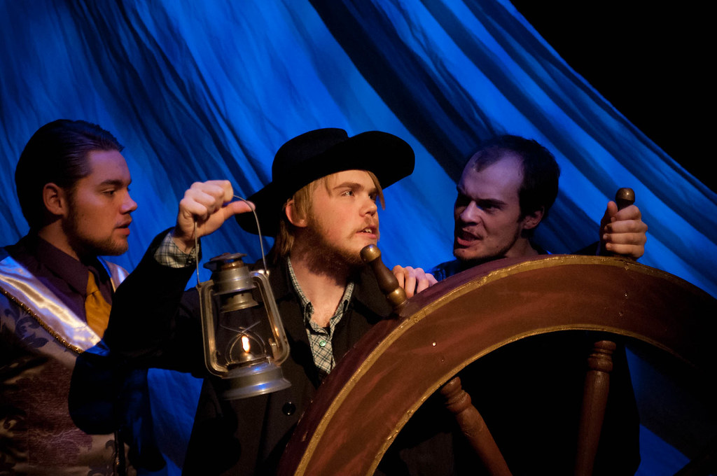 Three students at Bethany Lutheran College perform on stage under blue lighting: one holds a lantern while the group stands around a ship’s wheel, depicting a dramatic scene from a play.