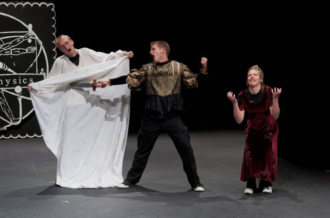 Three Bethany Lutheran College students perform on stage; one is draped in a white sheet, another wears a gold and black top, and the third, dressed in a red dress, makes dramatic gestures.