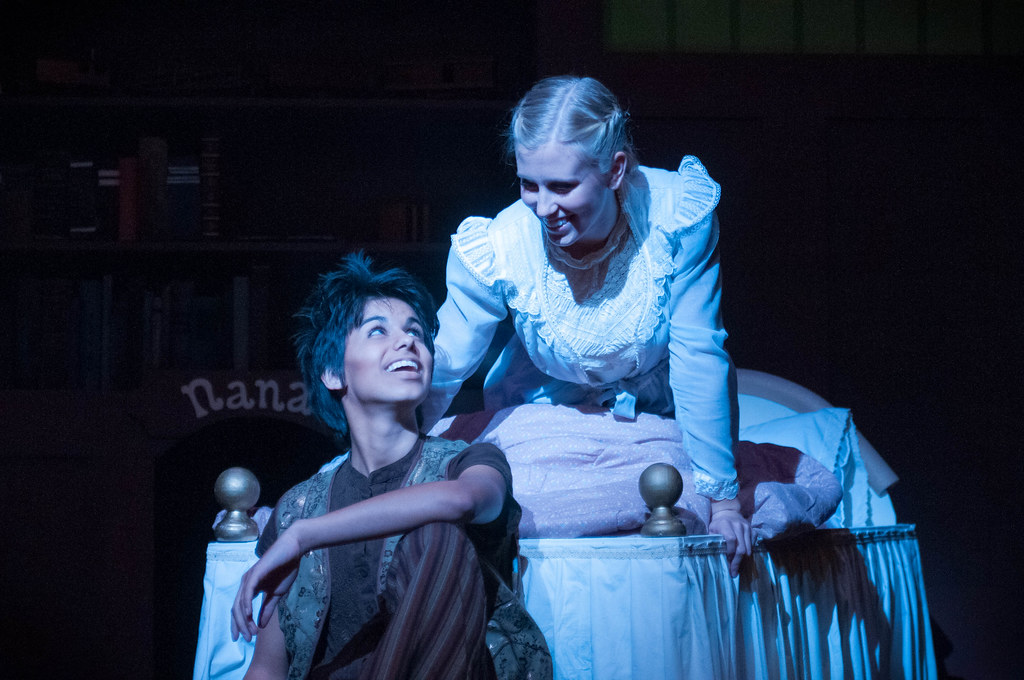 Two Bethany Lutheran College students perform in a stage production; one sits on the floor by a bed while the other kneels on the bed. Both are in costume, highlighted under stage lighting, suggesting an emotional or dramatic scene.