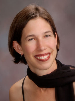 A woman with short brown hair, dressed in a black sleeveless top and a black scarf, smiles at the camera against a gray background. This image may be suitable for a professional profile photo for Bethany Lutheran College.