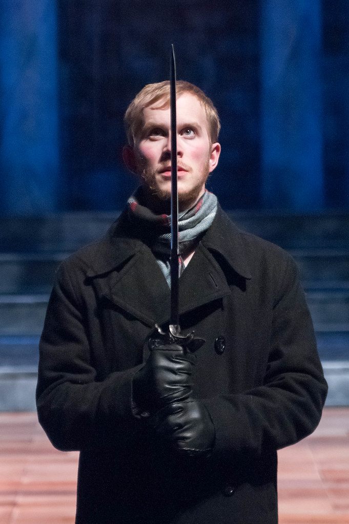 A man in a coat and gloves holds a sword upright in front of his face on a dimly lit stage, likely performing during a theatrical production at Bethany Lutheran College.