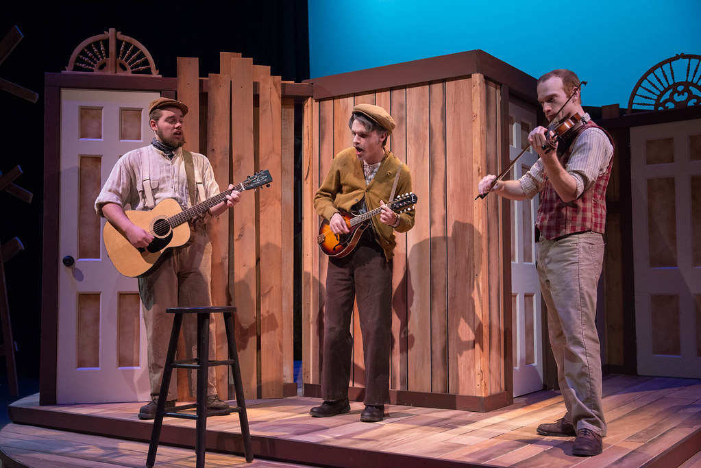 Three men perform on stage at Bethany Lutheran College, playing guitar, mandolin, and violin in front of wooden set pieces.