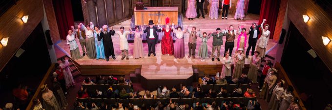 A theater cast in costume stands on stage at Bethany Lutheran College, holding hands for a curtain call as the audience of students and attendees watches from their seats below.
