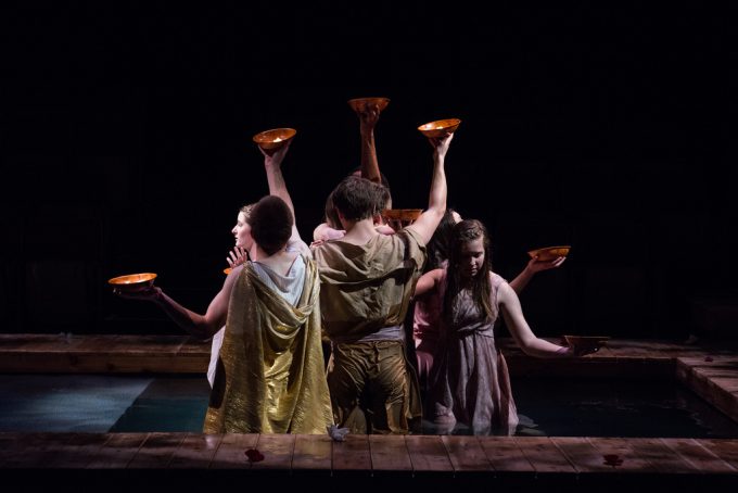 Four students in costumes stand close together, each holding a bowl aloft, on a dimly lit stage with a wooden border at Bethany Lutheran College.
