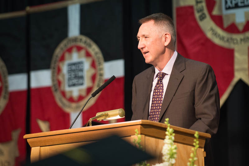 A man in a suit speaks at a podium during an indoor event at Bethany Lutheran College, with banners displayed in the background.