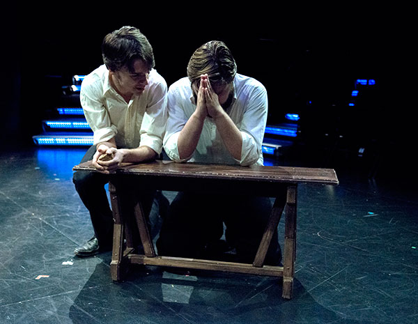 Two students in white shirts perform a scene on stage at Bethany Lutheran College, seated at a wooden bench; one student sits with their head in their hands while the other leans in to speak.