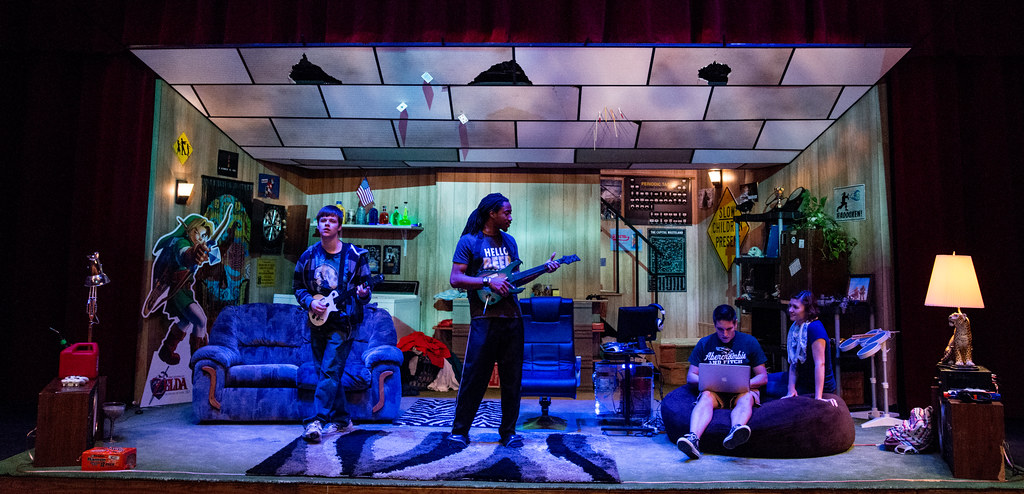 A stage set at Bethany Lutheran College portrays a living room scene with four students: two engaged in video games, one using a laptop, and another sitting on the floor.