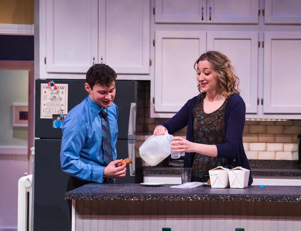 Bethany Lutheran College students enjoy a friendly moment in a modern kitchen, as a woman pours milk into a glass and a smiling man holds food beside her. White cabinets provide a bright, inviting backdrop to their interaction.