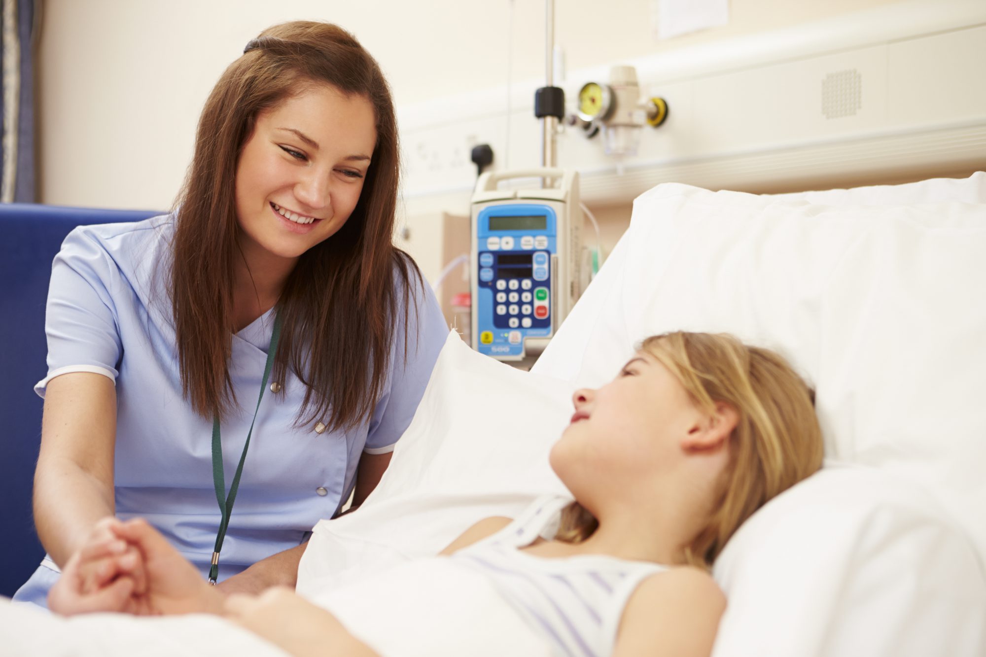 A nurse smiles and holds hands with a student lying in a hospital bed, with medical equipment visible in the background. (Bethany Lutheran College).