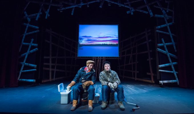 Two students sit on a stage at Bethany Lutheran College with fishing gear, while a winter scene is projected on the screen behind them.