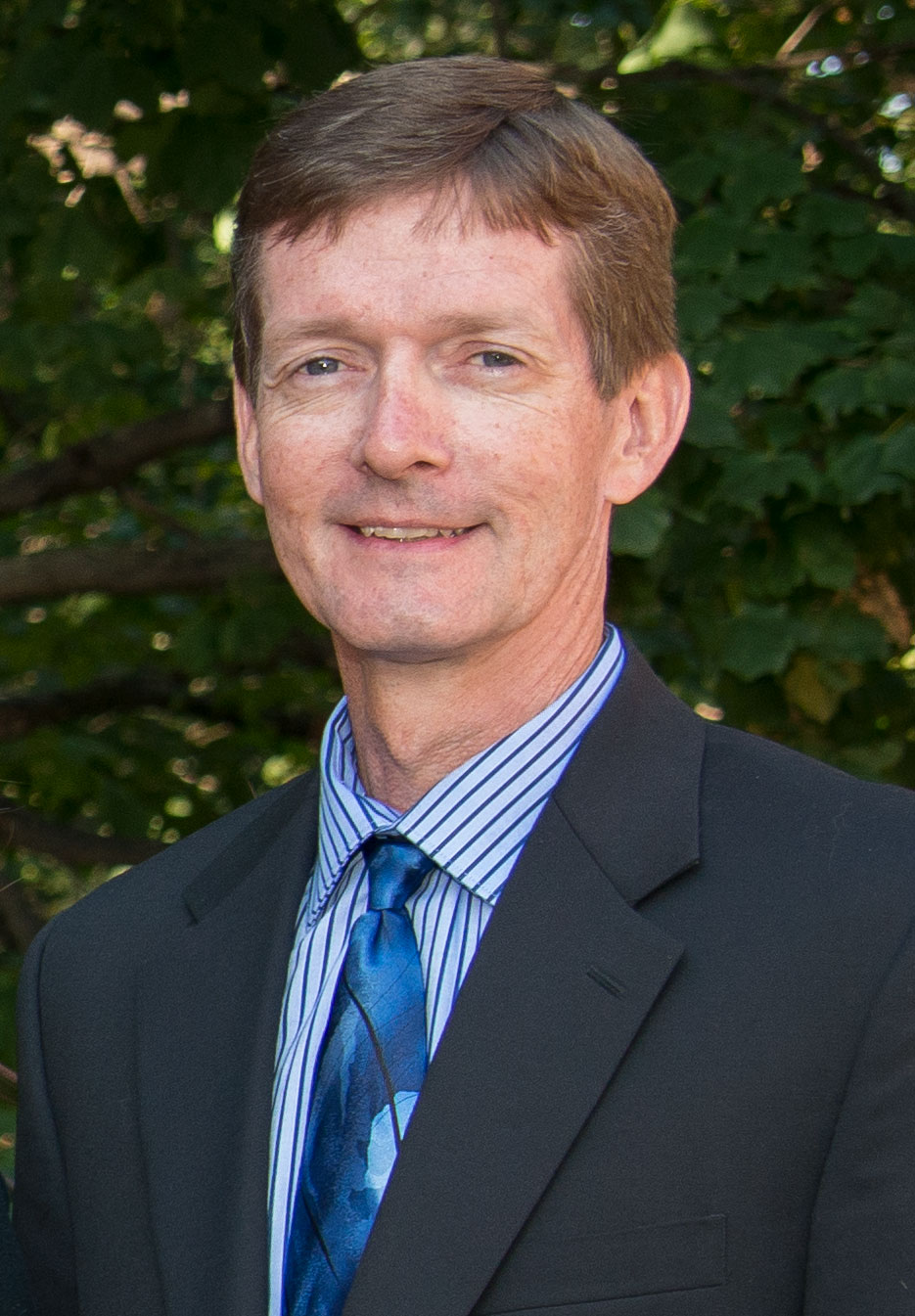 A man in a suit and striped shirt stands outdoors in front of green leafy trees, looking at the camera.