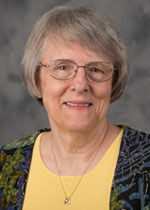 An older woman with short gray hair and glasses, wearing a yellow top and patterned jacket, smiles at the camera. She appears to be affiliated with Bethany Lutheran College.