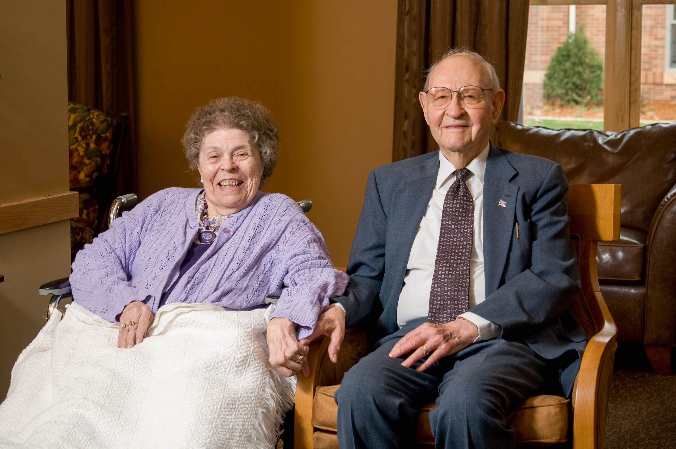 An elderly woman in a wheelchair and an elderly man in a suit sit side by side indoors, holding hands and smiling, possibly at a Bethany Lutheran College event or gathering.