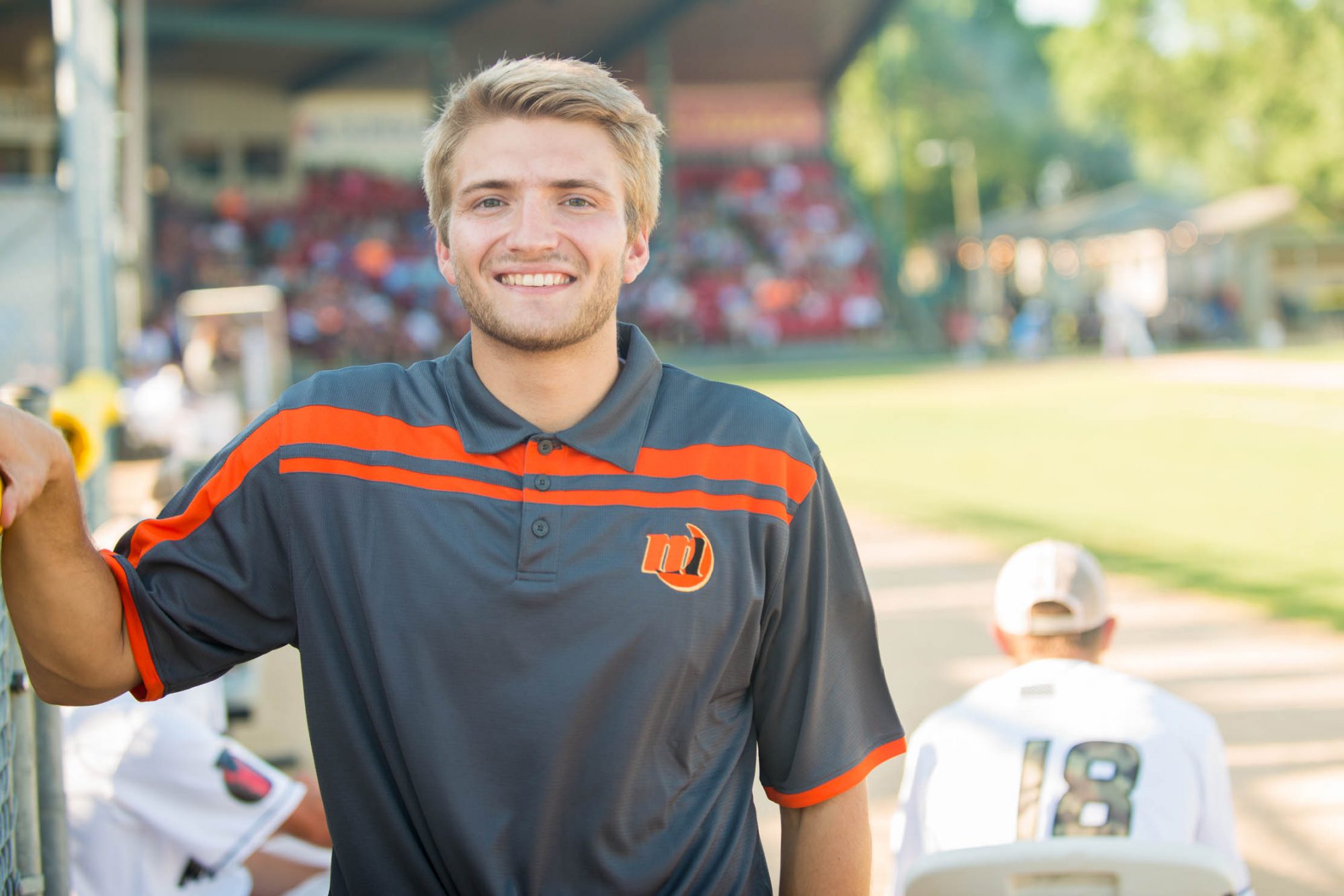 A young man in a dark gray and orange polo shirt stands and smiles at a baseball field, with players visible in the background. This image may be related to Bethany Lutheran College athletics or student activities if taken on their campus.