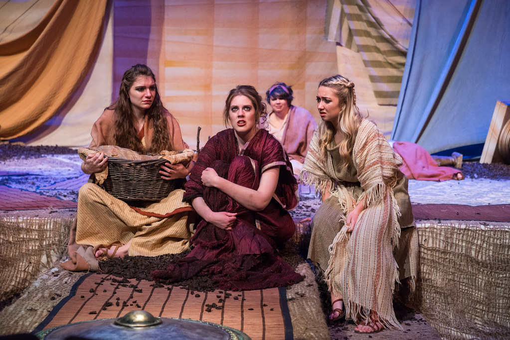 Three students from Bethany Lutheran College, dressed in robes, sit on stage with draped fabric as a backdrop; one holds a basket while another is seated behind them.