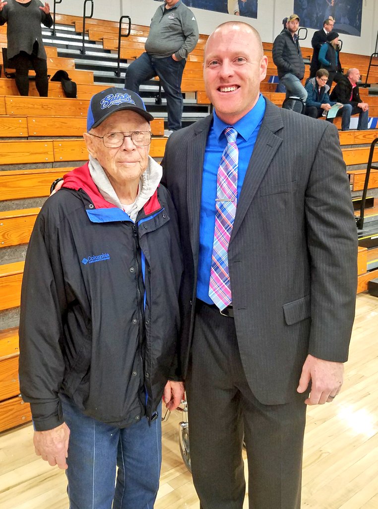 An older man in casual attire stands beside a man in a suit inside the Bethany Lutheran College gymnasium, with wooden bleachers visible in the background.