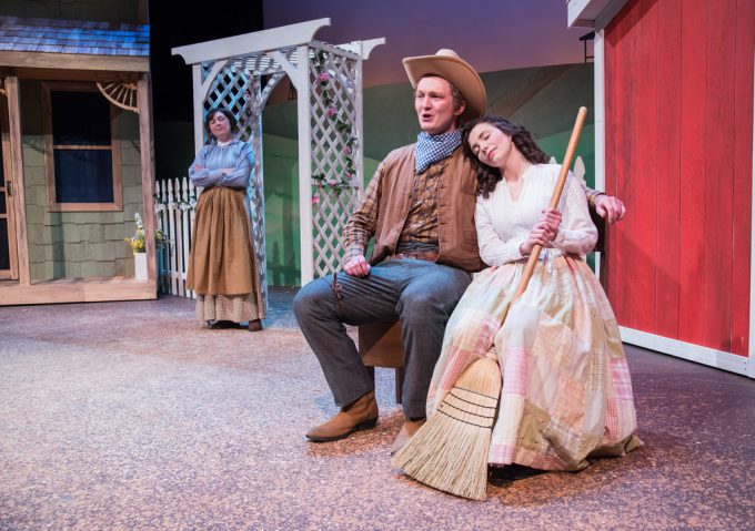 Three actors, likely Bethany Lutheran College students, perform on stage in period costumes. A man and a woman are seated together while another woman stands in the background, suggesting a dramatic scene from a historical or classical play.