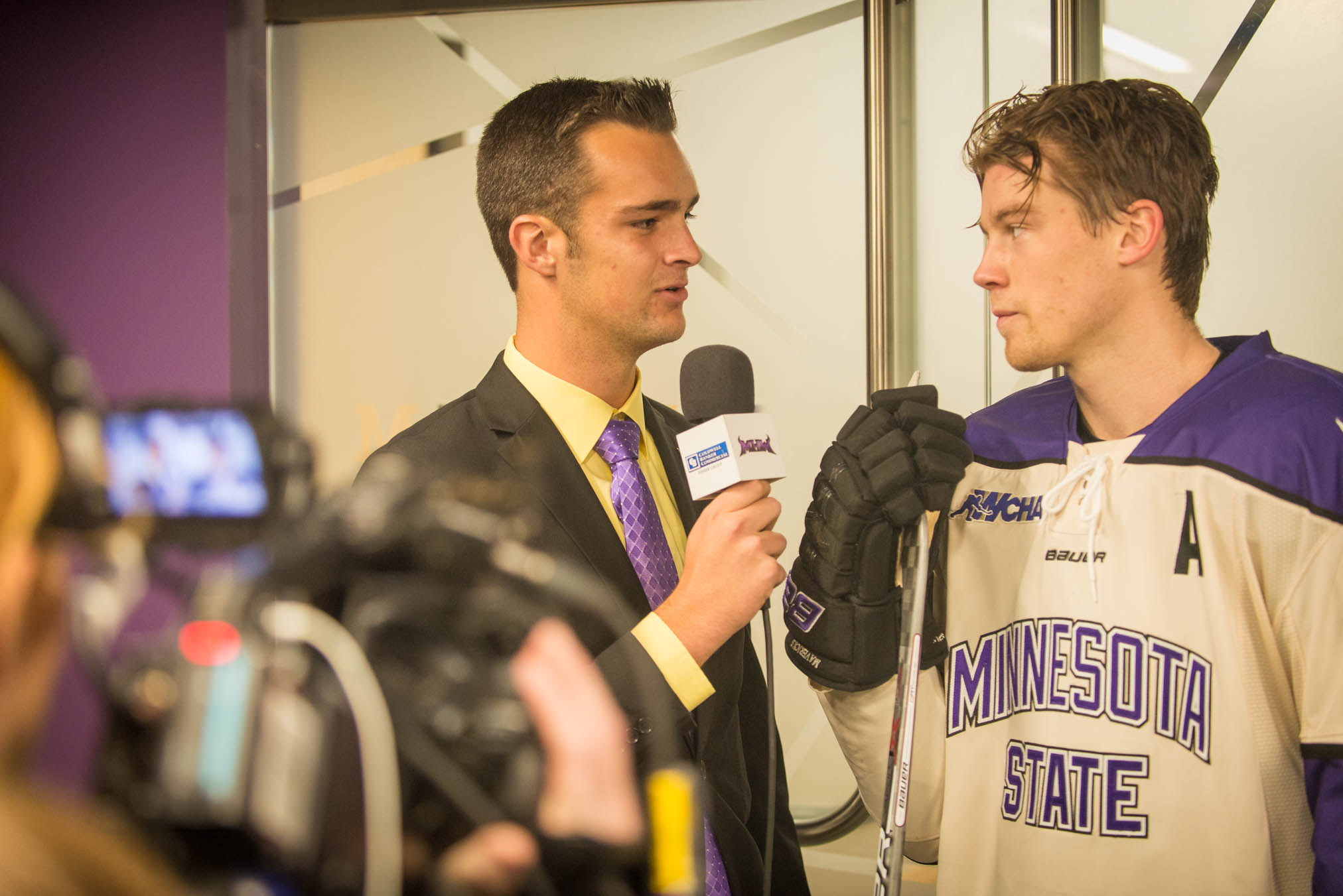 A reporter interviews a hockey player wearing a Minnesota State jersey, while a camera crew films the exchange. If this event is related to Bethany Lutheran College, it could be covering student athletes or college sports representation.