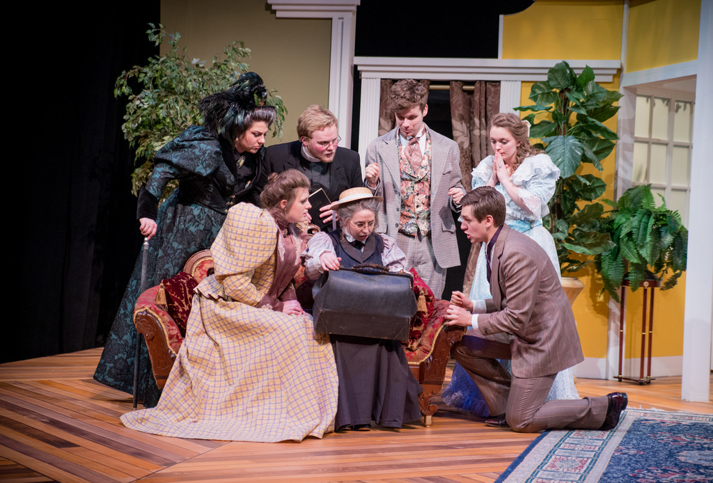 Seven people in period costumes gather closely around a seated woman opening a black bag on a stage set, likely during a theatrical performance at Bethany Lutheran College.