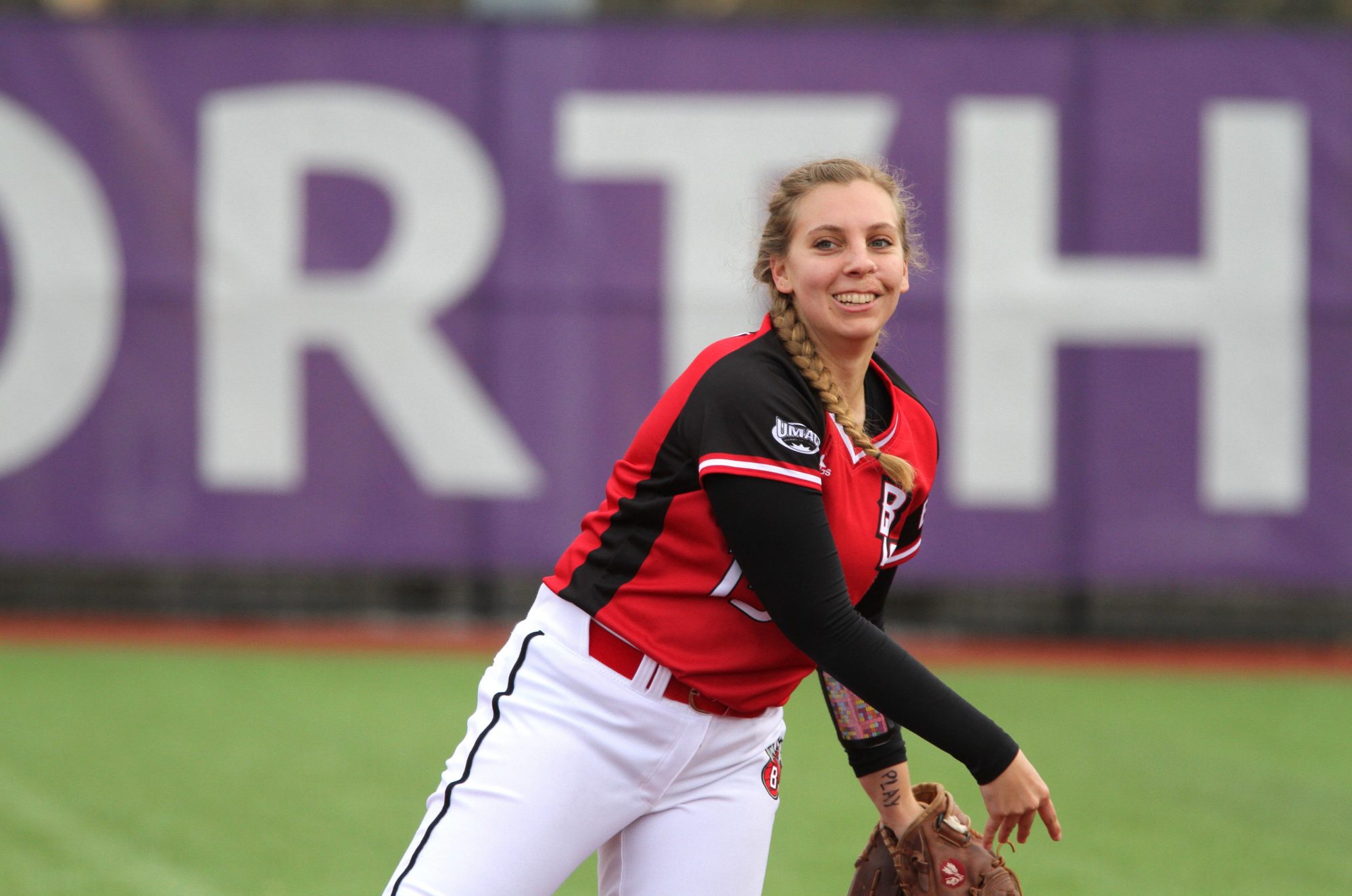 A Bethany Lutheran College softball player in a red and black uniform stands on the field, smiling with a glove on her left hand.