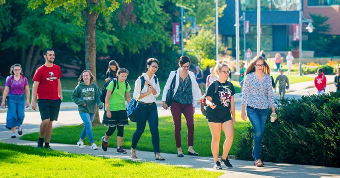 A group of Bethany Lutheran College students walks together on a sunny campus sidewalk, surrounded by green grass and trees.
