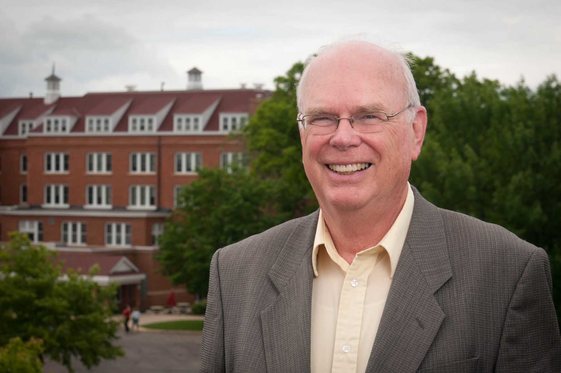 An older man in a suit jacket and glasses stands outside in front of a large brick building with trees nearby, possibly on the campus of Bethany Lutheran College.