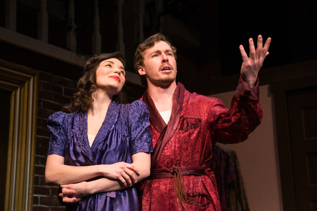Two students in theatrical costumes stand indoors, performing a scene. The woman in a purple dress holds the mans arm as he gestures upward. This is likely from a Bethany Lutheran College theater production.