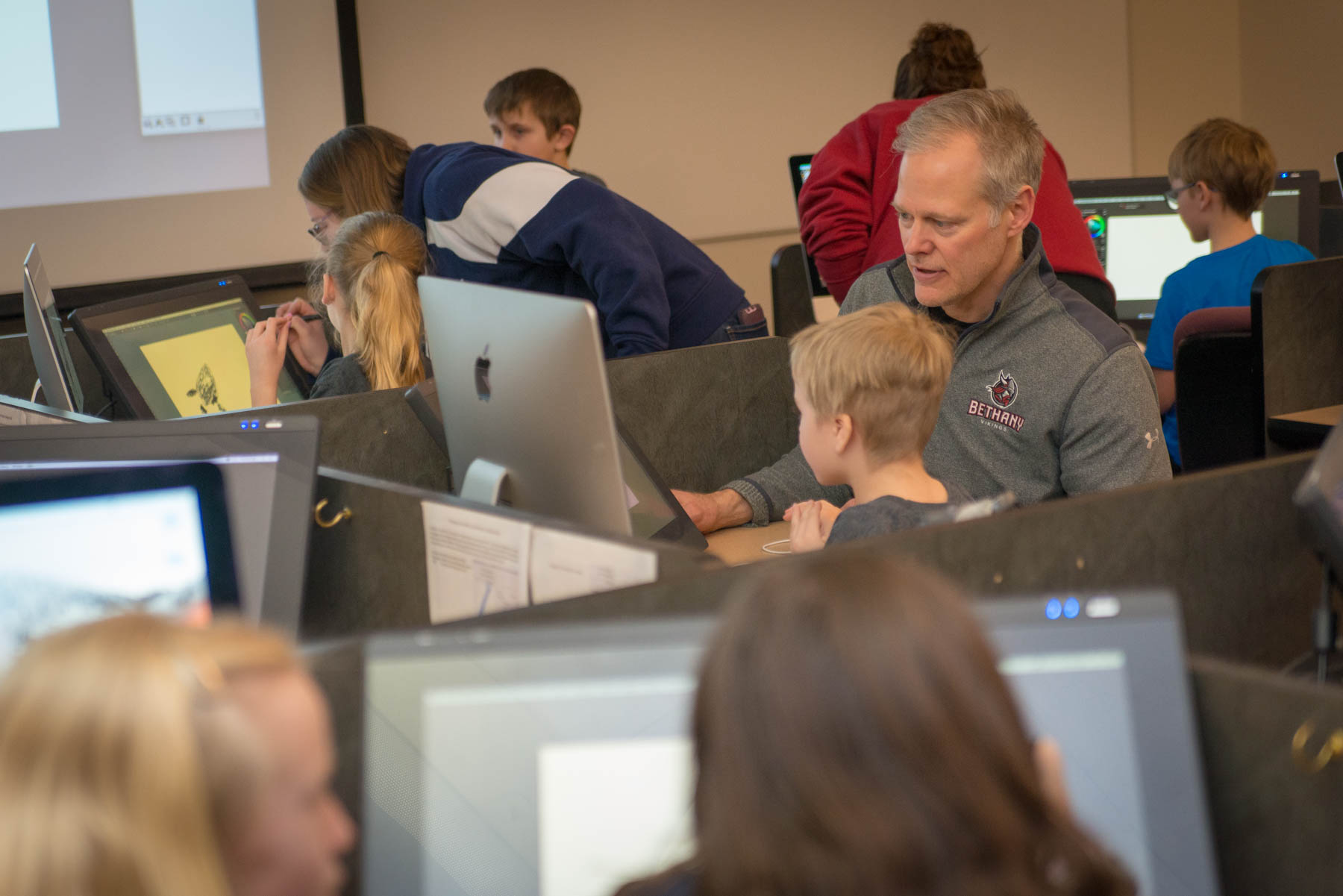 Students and adults work together on computers in a Bethany Lutheran College classroom, as a man assists a young boy at his monitor.