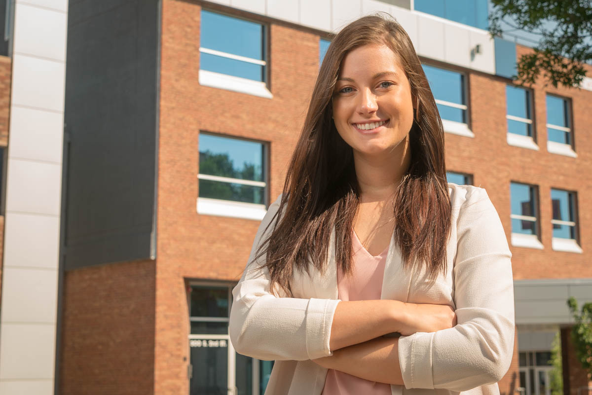 A woman with long brown hair stands outside a modern brick building at Bethany Lutheran College, smiling confidently with her arms crossed.