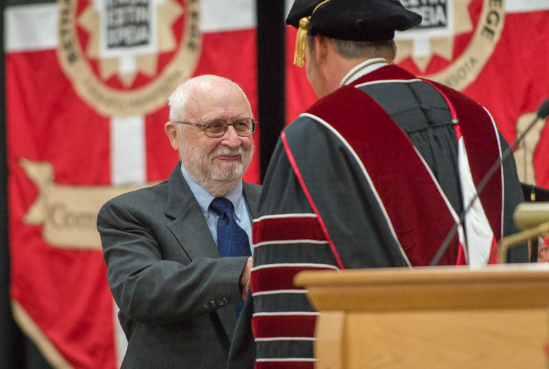 An older man in a suit shakes hands with a person in academic regalia during a ceremony at Bethany Lutheran College, with red banners visible in the background.