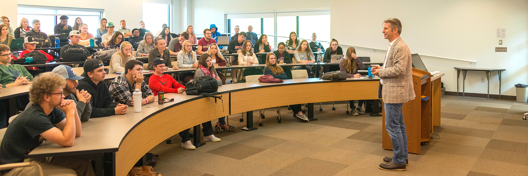 A Bethany Lutheran College professor stands at a podium delivering a lecture to students seated in a curved lecture hall classroom.