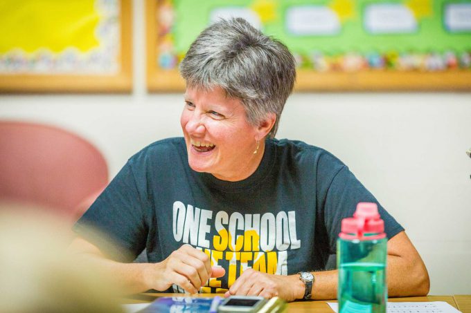 A smiling individual with short gray hair is seated at a table, wearing a black One School, One Team t-shirt. This image may depict a staff or faculty member in a casual setting at Bethany Lutheran College. No students are present.
