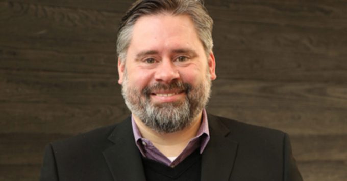 A bearded man wearing a black blazer and collared shirt stands in front of a dark wooden wall.
