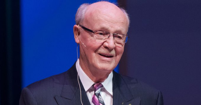 An older man in a suit, tie, and glasses speaks while wearing a headset microphone against a blue background, likely delivering a lecture or presentation at Bethany Lutheran College.