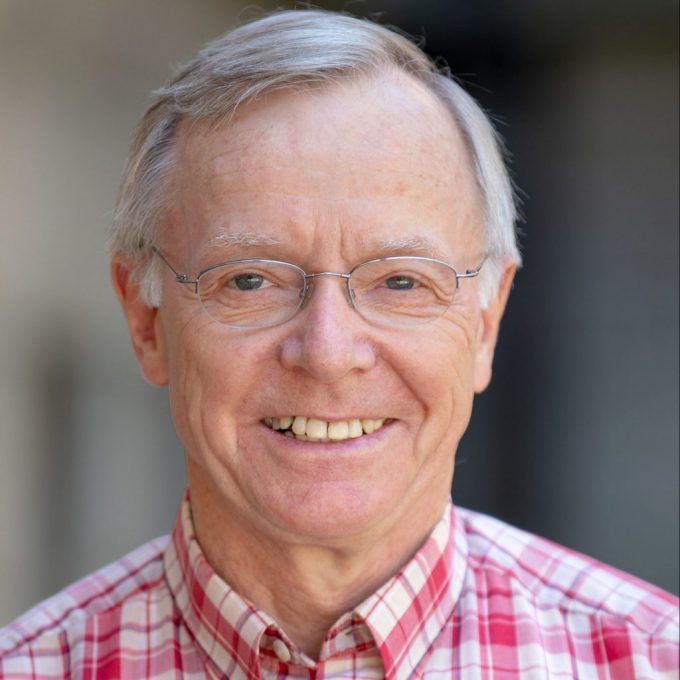 An older man with glasses and gray hair smiles while wearing a red and white plaid shirt. The background is blurred, focusing attention on the subject.
