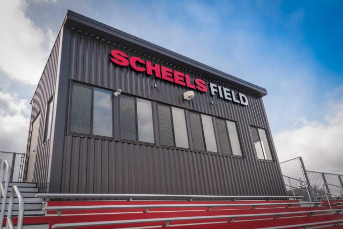 The photo shows the Bethany Lutheran College SCHEELS Field press box, a black structure with large windows, situated above red stadium seats and prominently displaying the red SCHEELS FIELD sign against a blue sky.