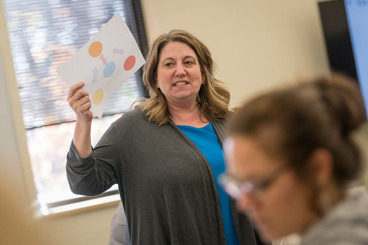 A woman presents a paper with colored shapes to a group of students during a classroom or meeting at Bethany Lutheran College.