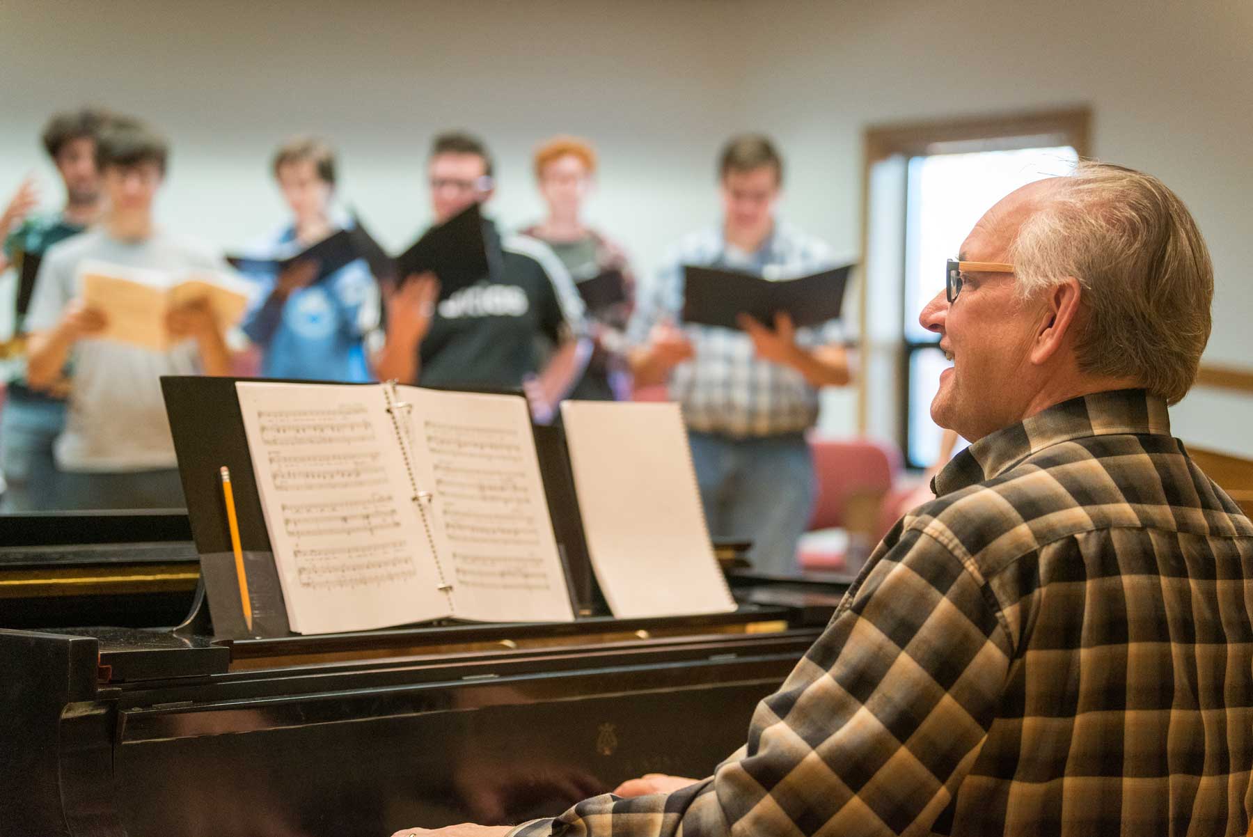 An older man plays piano while Bethany Lutheran College students sing and hold open music books in a bright room.