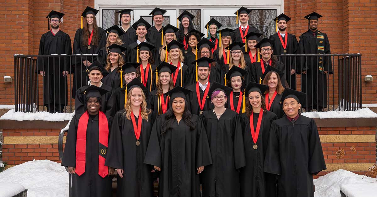 A group of graduates from Bethany Lutheran College in caps and gowns stand on the steps outside a brick building, posing for a group photo.