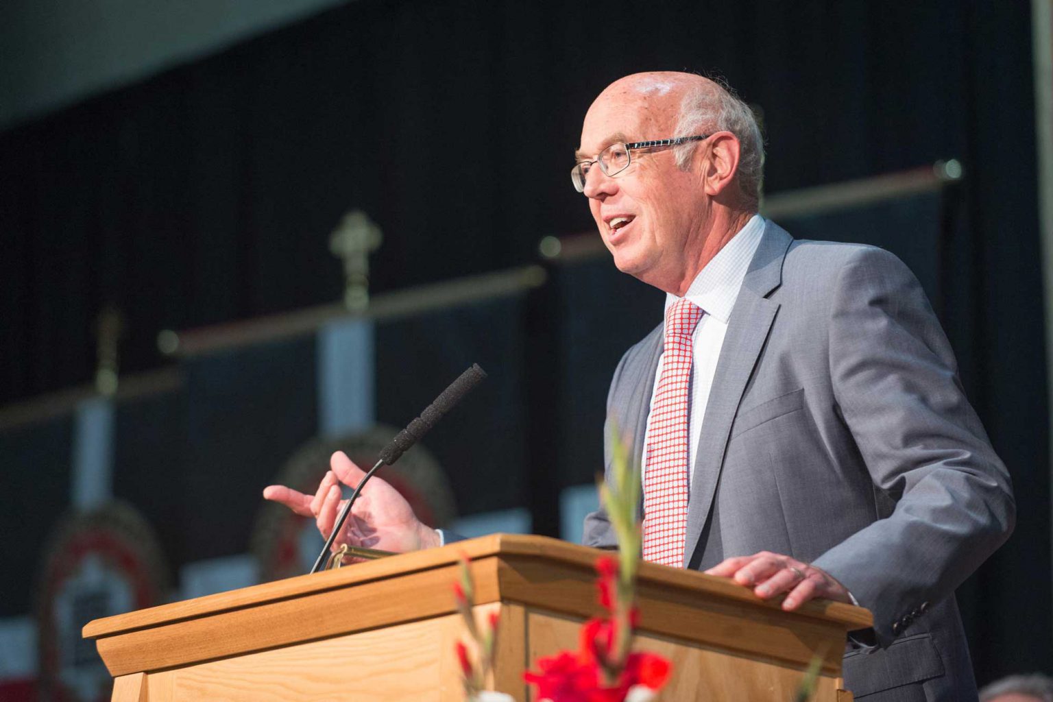 A man in a suit and tie speaks at a podium with a microphone, with banners in the background and flowers arranged in the foreground at Bethany Lutheran College.
