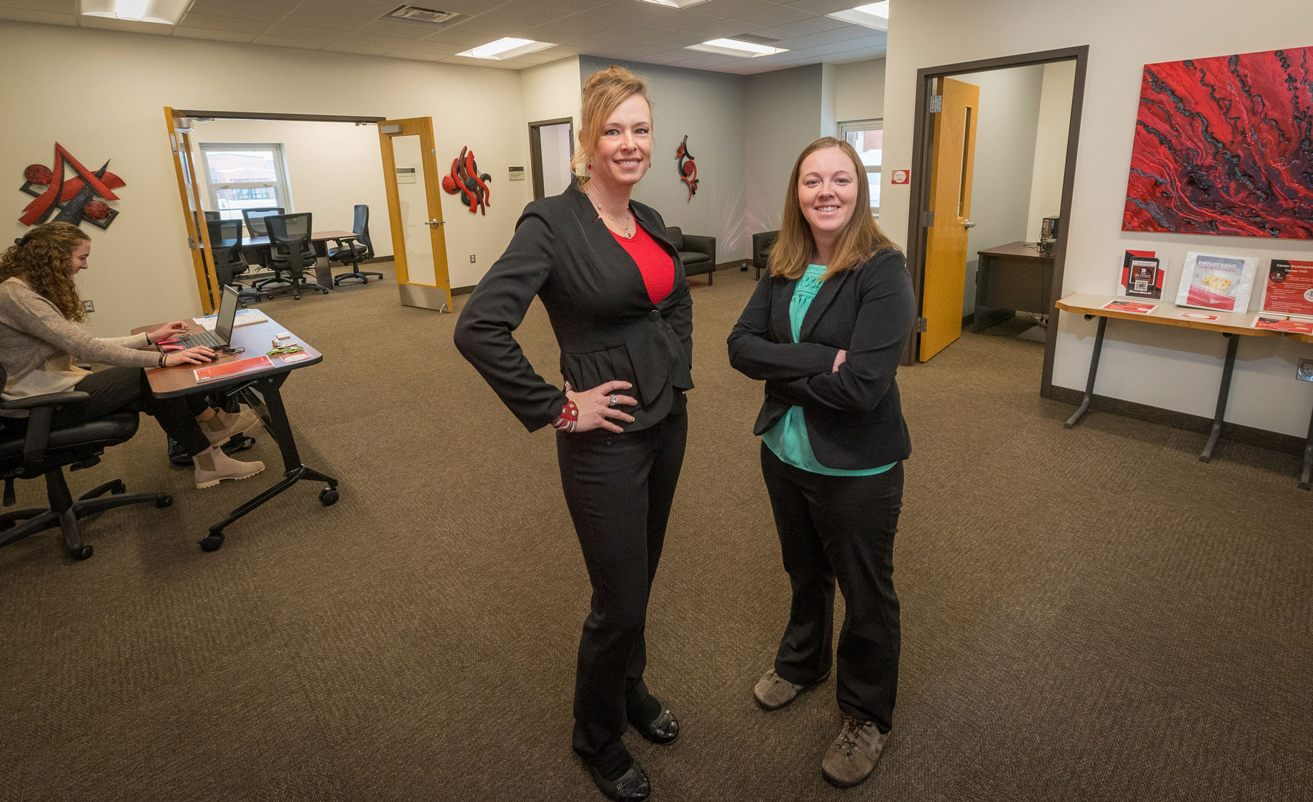 Two women in business attire stand in an office space at Bethany Lutheran College, with another person working at a desk in the background.