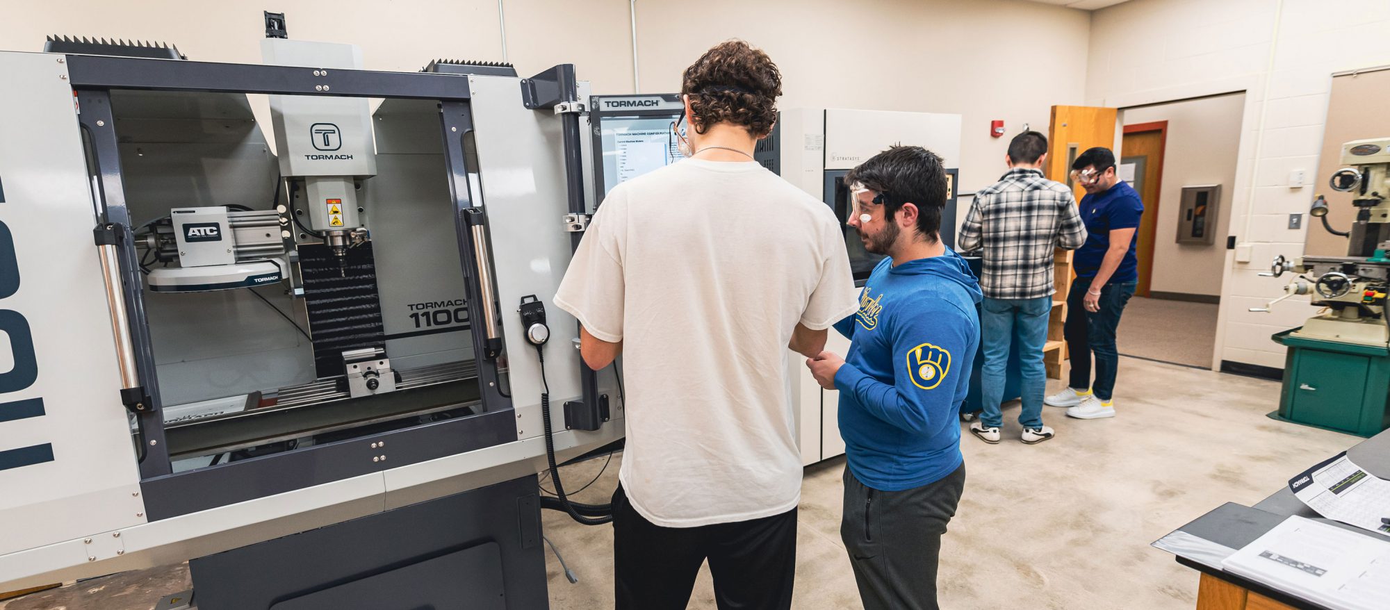 Students at Bethany Lutheran College work on and observe machines in a workshop or lab equipped with industrial equipment and computers.