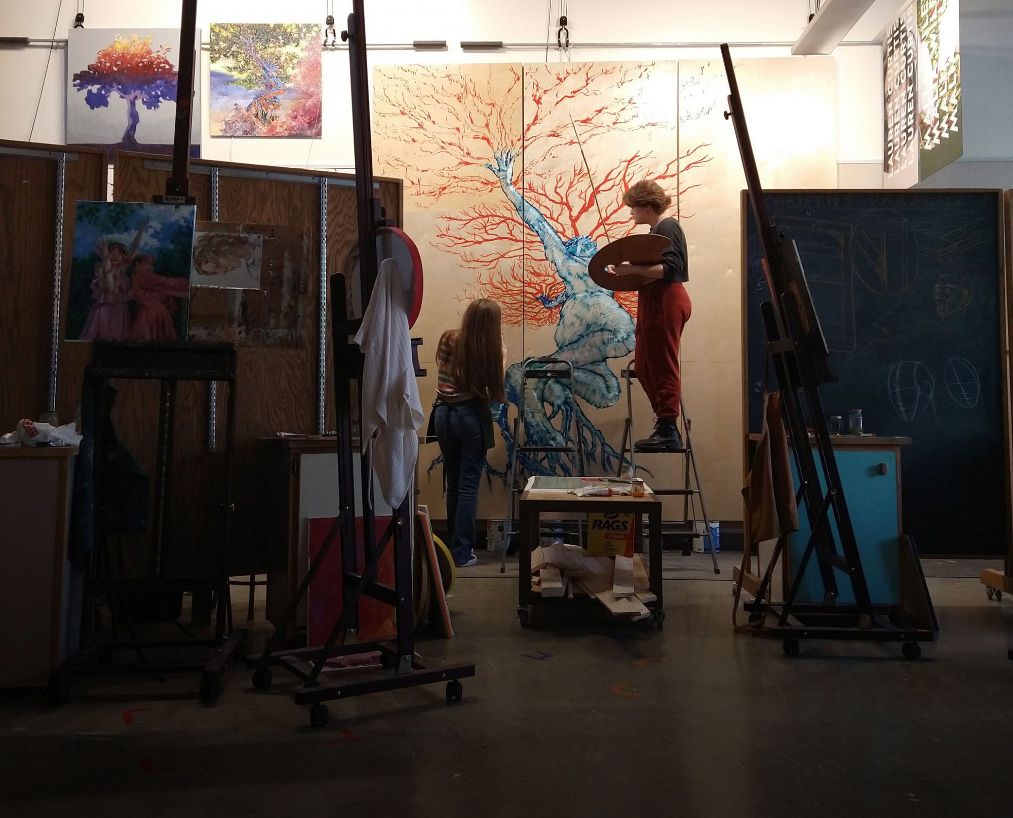 Two Bethany Lutheran College students collaborate on a large tree painting in an art studio, surrounded by easels and various art supplies.