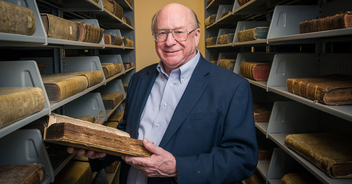 An older man in a suit jacket stands between shelves filled with large, antique books at Bethany Lutheran College, holding an old volume, likely showcasing the colleges rare book collection or library resources.