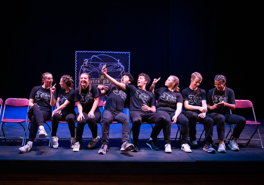 Eight students in matching black t-shirts sit on stage chairs at Bethany Lutheran College; some pose for a group selfie while others look around.