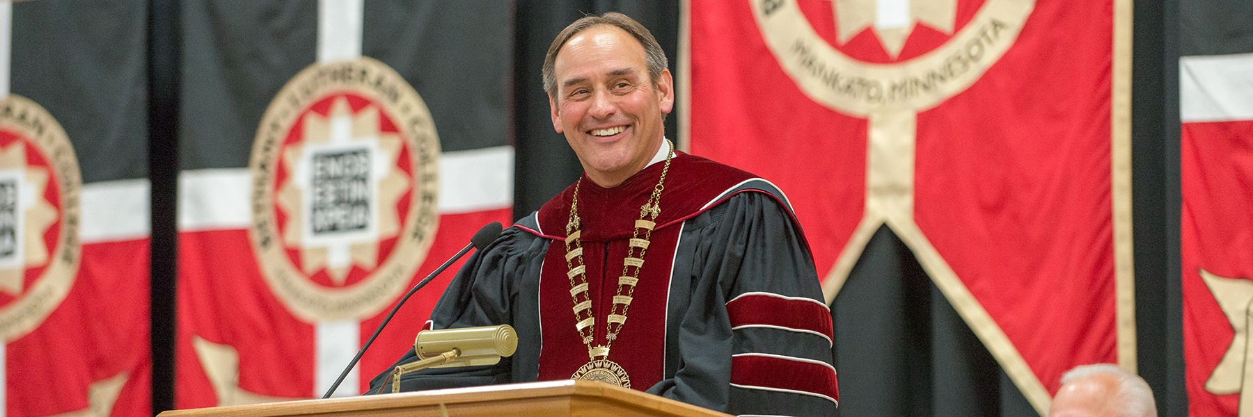 A man in academic regalia speaks at a podium adorned with red and black banners in the background during a formal event at Bethany Lutheran College.