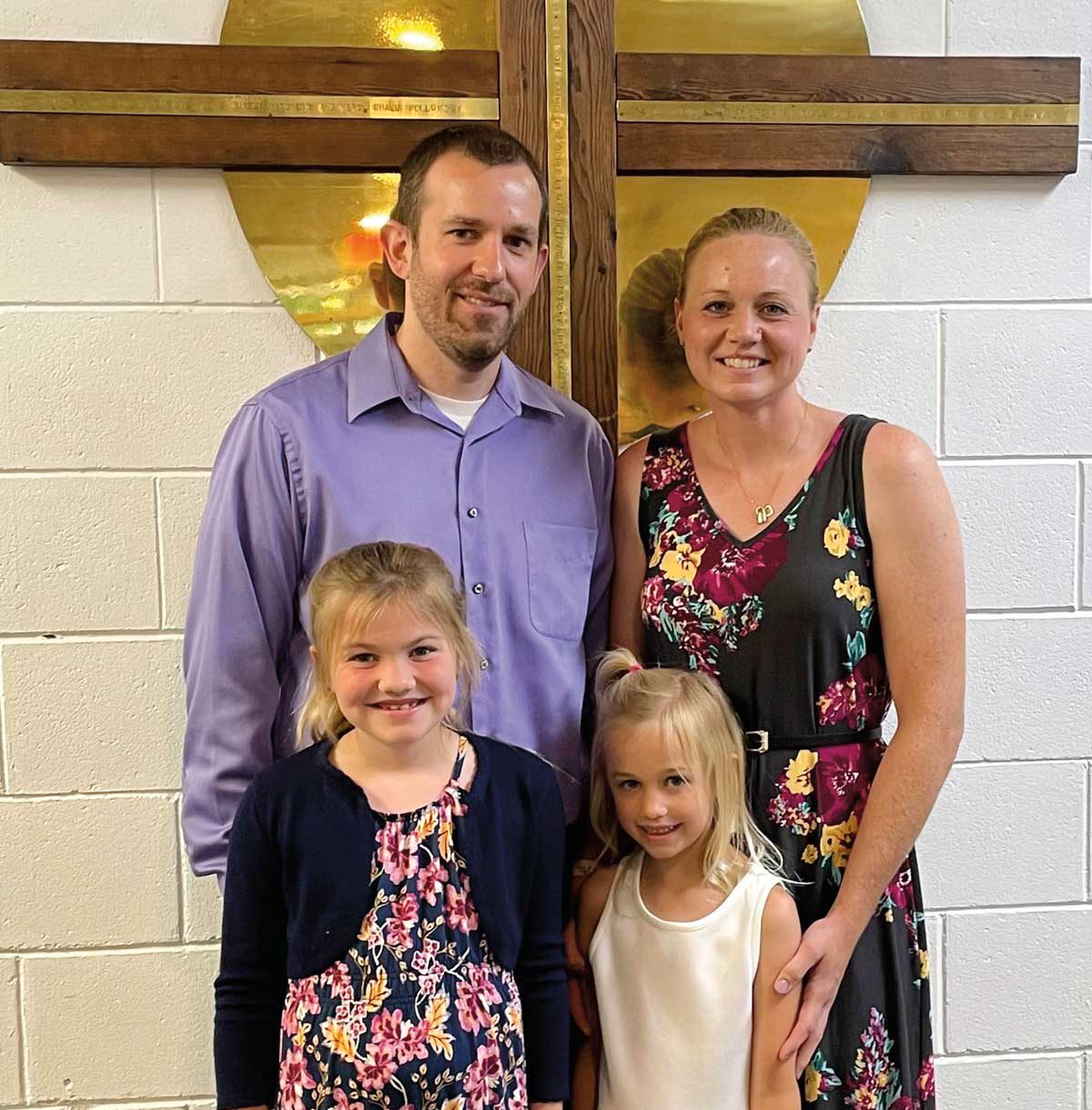 A man and woman stand behind two students in front of a cross mounted on a white brick wall, possibly at Bethany Lutheran College.