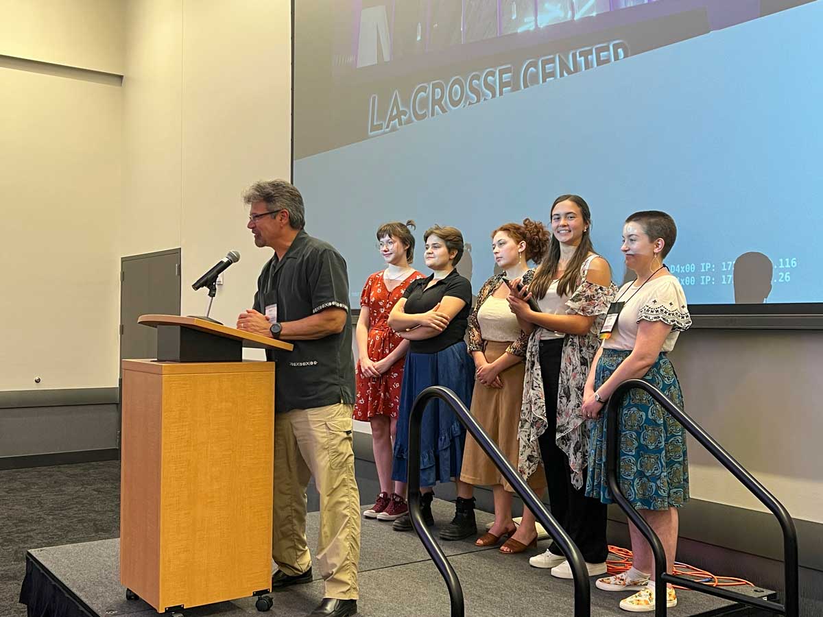 A man speaks at a podium while six women stand in a line on stage in front of a projection screen at Bethany Lutheran College.